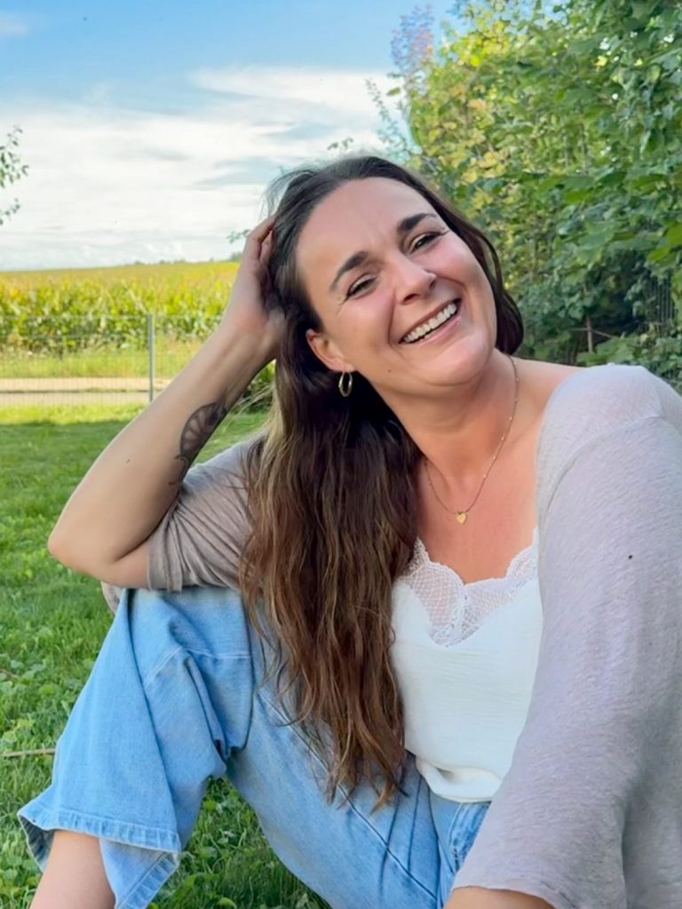 Melanie Schmid - Soul Osteopathie Wolfersdorf-Oberhaindlfing Junge Frau sitzt lächelnd im Grünen, umgeben von Bäumen und einem klaren Himmel.